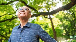 An older Asian man in a blue plaid shirt and glasses looks up while standing among green trees.