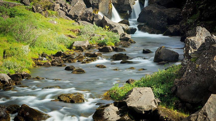 A smooth, milky river flows through a rocky landscape with green banks and a small waterfall in Thingvellir, Iceland.