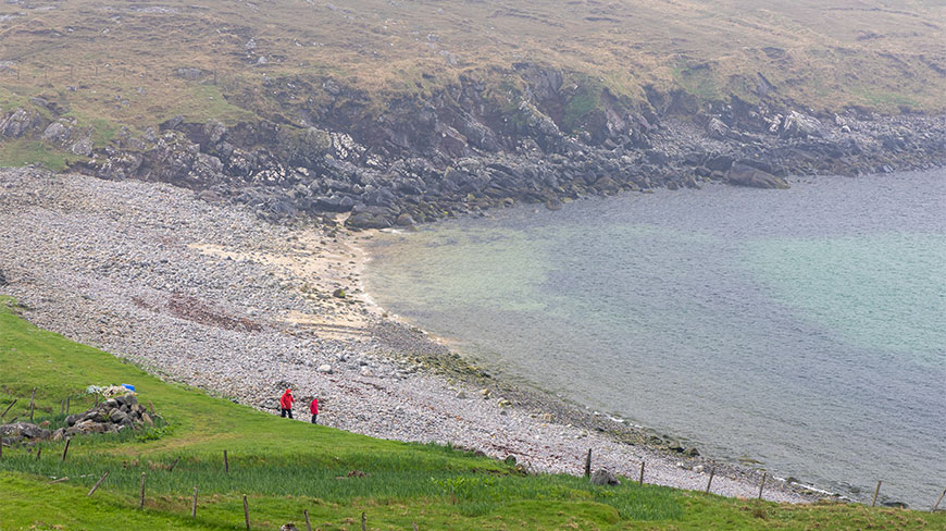 Two people in red jackets walk along a path above a pebble beach on the misty coast of Stornoway, Scotland.