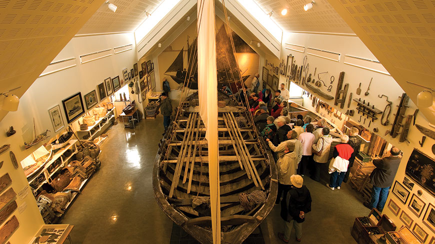A group of visitors observes a large wooden boat on display inside the Skogar Museum in Iceland, with various artifacts lining the walls.