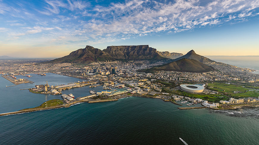 A stunning aerial view of Cape Town's city bowl and harbor, with the iconic Table Mountain prominent against a partly cloudy sky.