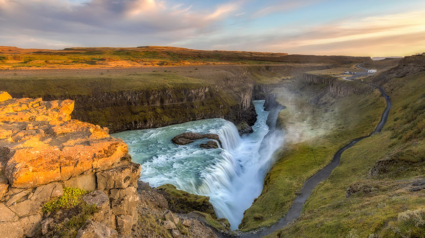 The powerful Gullfoss Falls cascades through a vast, green canyon in Iceland under a colorful sky at sunset.