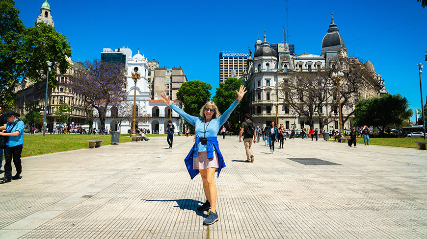 A smiling woman with arms raised stands in a sunny city square surrounded by historic buildings and other people.