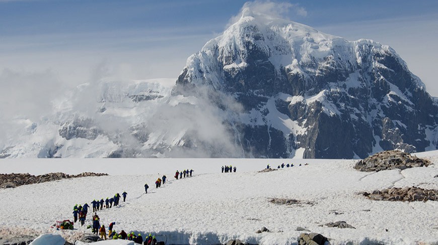 A group of explorers hike up a snowy Antarctic peninsula with a towering, cloud-wreathed mountain in the background under a blue sky.