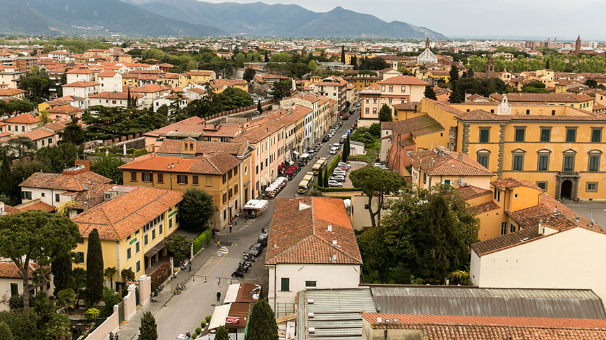 An aerial view of the city of Pisa, Italy, with its terra-cotta-roofed buildings and mountains rising in the background.