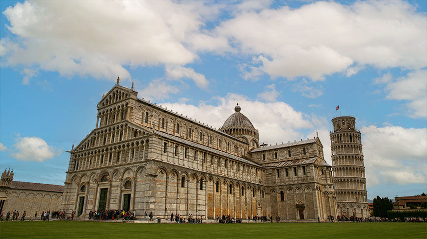 The Leaning Tower of Pisa and Pisa Cathedral on a green lawn under a blue sky with fluffy clouds in Italy.