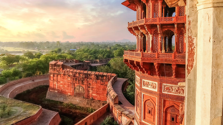 The sun sets over the red sandstone Agra Fort in India, illuminating its ornate walls and the lush landscape beyond.