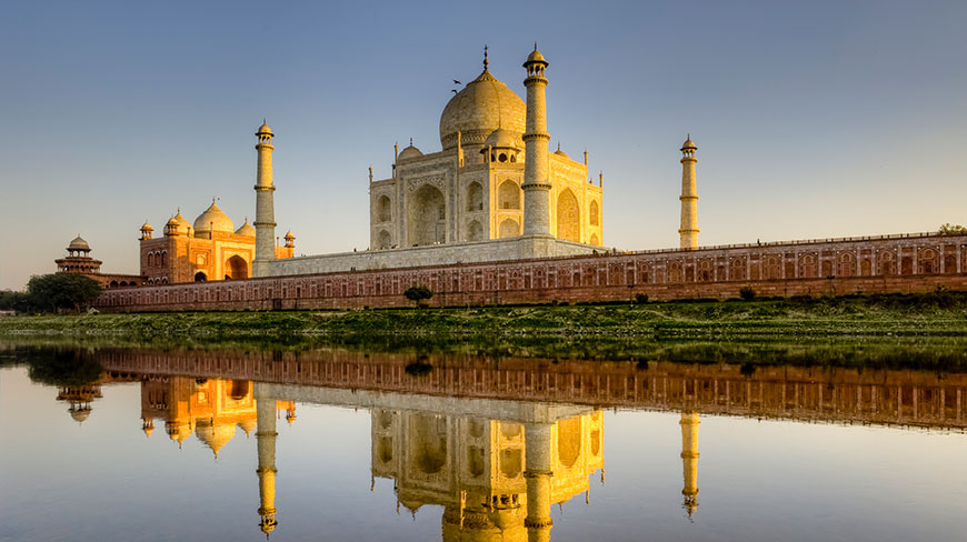 The Taj Mahal in India is bathed in golden morning light, with its perfect reflection visible in the calm river below.