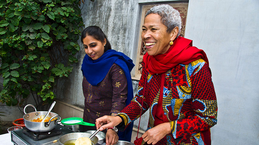 Two women participate in an outdoor cooking demonstration in India, smiling as one prepares a dish on a portable stove.