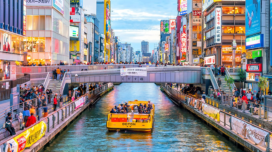 A yellow tour boat cruises down the Dotonbori canal in Osaka, Japan, surrounded by bustling crowds and vibrant buildings with colorful signs.