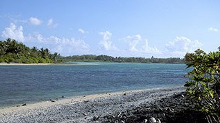 A rocky shoreline meets a turquoise lagoon with a lush, green island in the distance under a blue sky with scattered clouds.