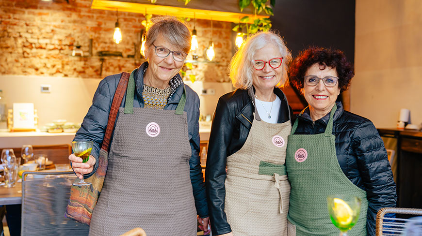 Three smiling women wearing aprons pose for a photo in a restaurant, two of them holding cocktails.