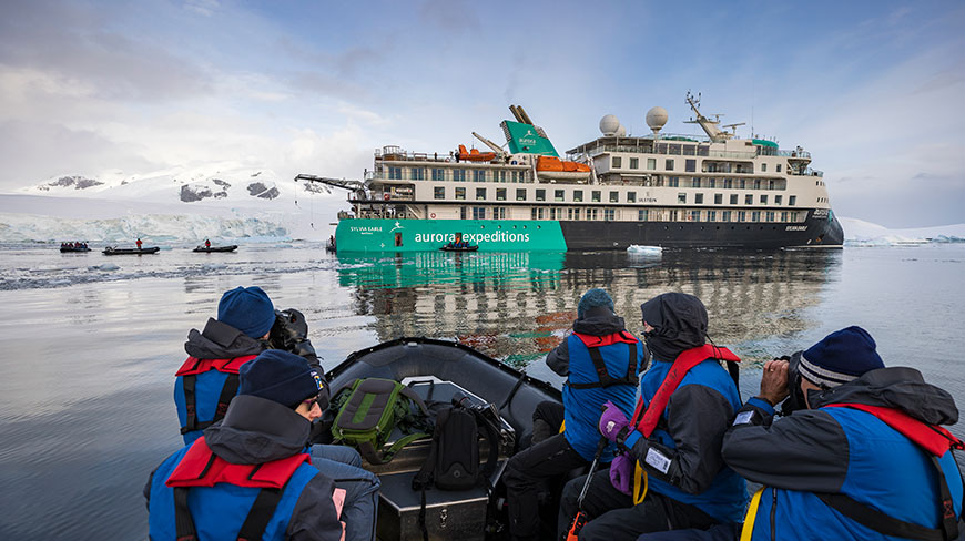 Passengers in a zodiac boat watch the large Sylvia Earle expedition ship with a snowy coastline in the background.