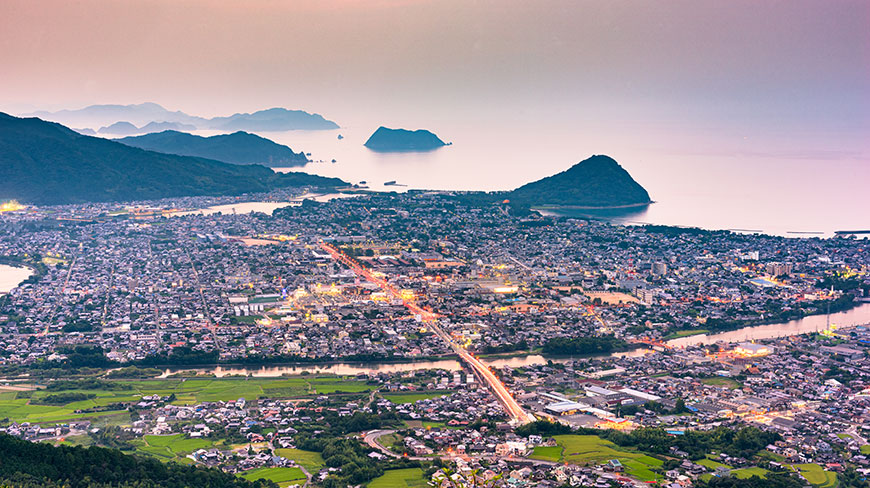 Aerial view of the coastal city of Hagi, Japan, at dusk, with city lights illuminating the streets and mountains in the background.