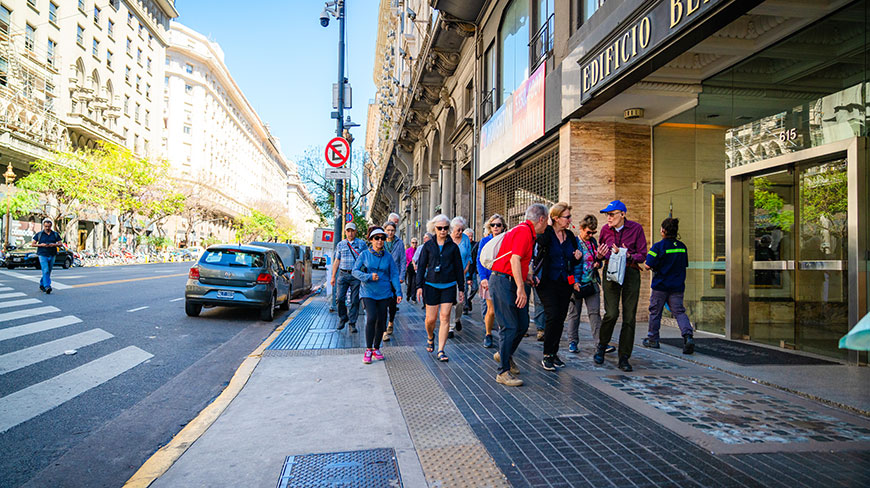 A group of travelers on a walking tour strolls down a wide city sidewalk in Buenos Aires on a sunny day.