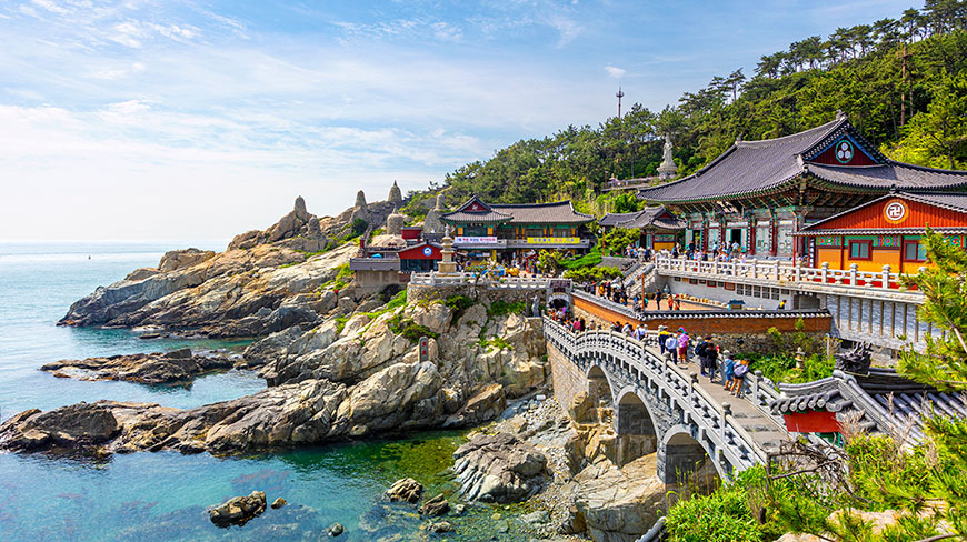 A vibrant temple complex perches on a rocky coast in Busan, South Korea, as visitors cross a stone bridge to the entrance.