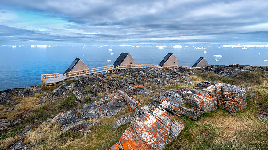 Four modern cabins on a rocky shore overlook the Ilulissat Icefjord, where icebergs float on the water in Greenland under a cloudy sky.