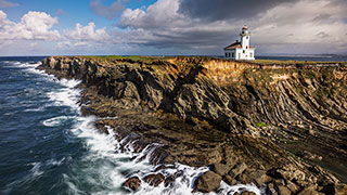 An aerial view of a white lighthouse on a rocky cliffside as waves from the ocean crash against the shore under a cloudy sky.