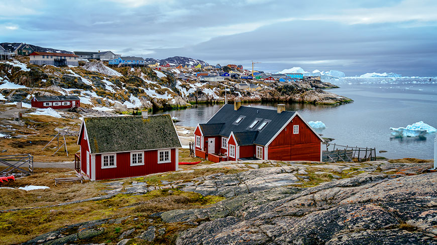 Colorful houses dot the rocky, snow-dusted coastline of Disko Bay, Greenland, with icebergs floating in the water.