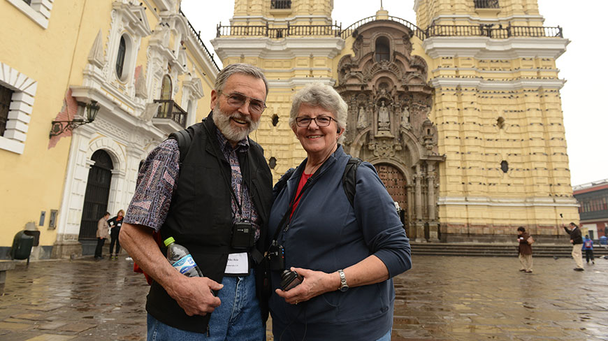A smiling couple stands in a plaza in front of an ornate, historic yellow church in Lima, Peru.