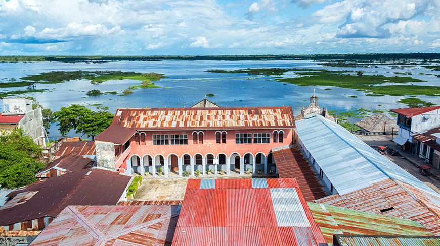 High-angle view of the city of Iquitos, Peru, showing rusty tin roofs and the vast, green-mottled Amazon River under a blue, cloudy sky.