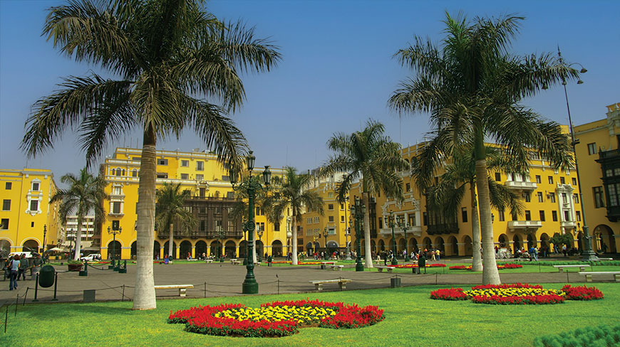 Palm trees stand on a grassy lawn in the historic Plaza de Armas in Lima, Peru, with yellow colonial buildings in the background.
