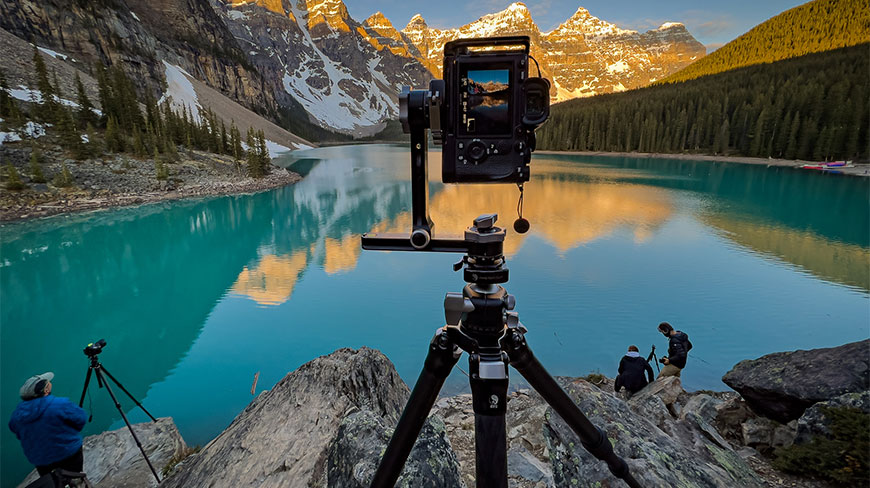 Photographers on a rocky overlook use tripods to capture the sunrise over a vibrant turquoise mountain lake.