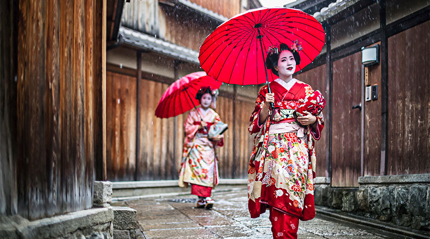 Two geishas in colorful kimonos hold red umbrellas while walking down a traditional, wet stone street in Japan.