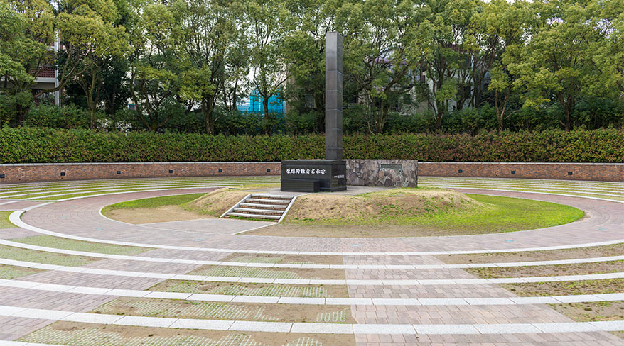 A tall, dark monument stands in a circular plaza at the Nagasaki Memorial in Japan, surrounded by lush green trees and manicured hedges.