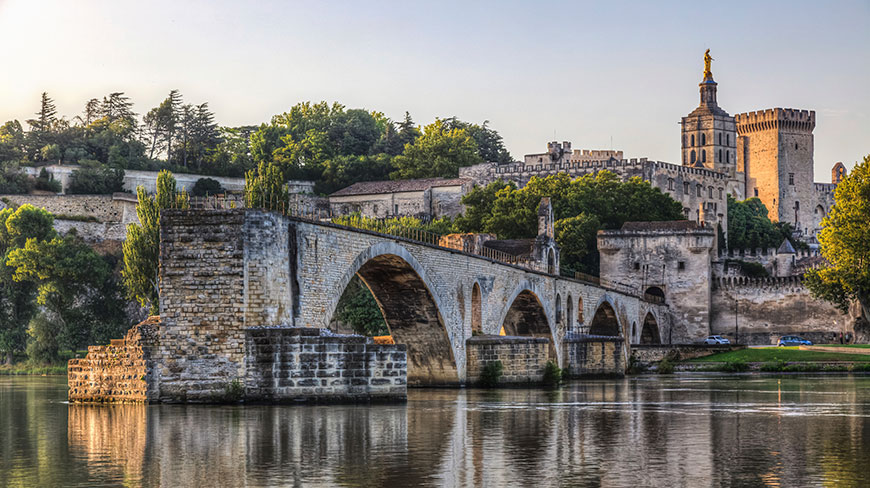 The historic Pont Saint-Bénézet arches over the Rhône river in Avignon, France, with the Palais des Papes in the background at sunset.